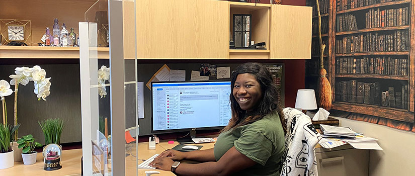 A woman with dark skin and dark hair sits at a computer. She is facing the camera and smiling. 