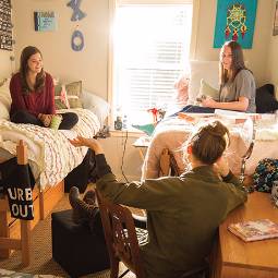 Three girls chatting in a dorm room, two sitting on beds and one seated at a desk, with sun shining in through the window blinds