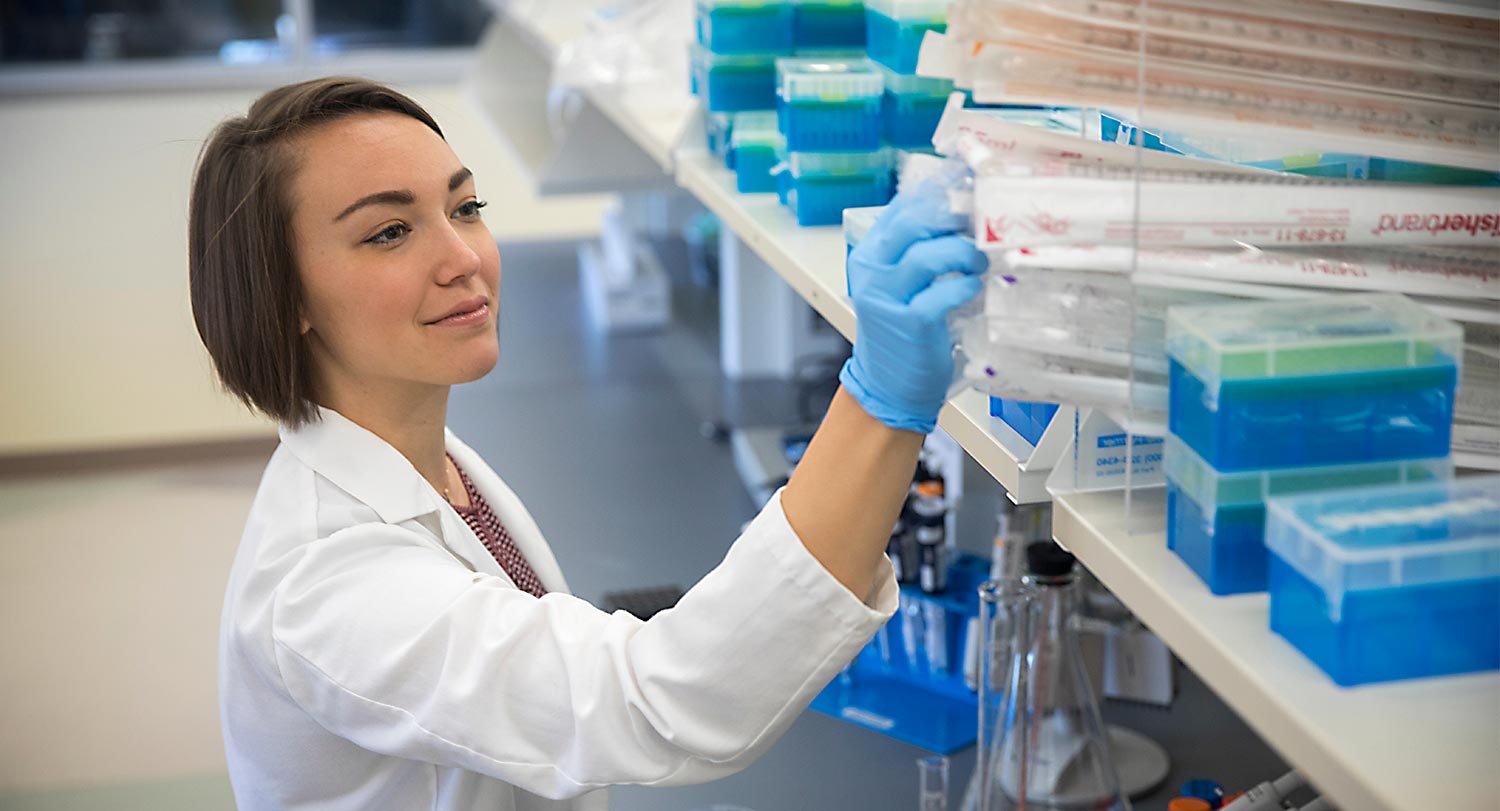 Researcher wearing a lab coat reaching for equipment on a shelf. 