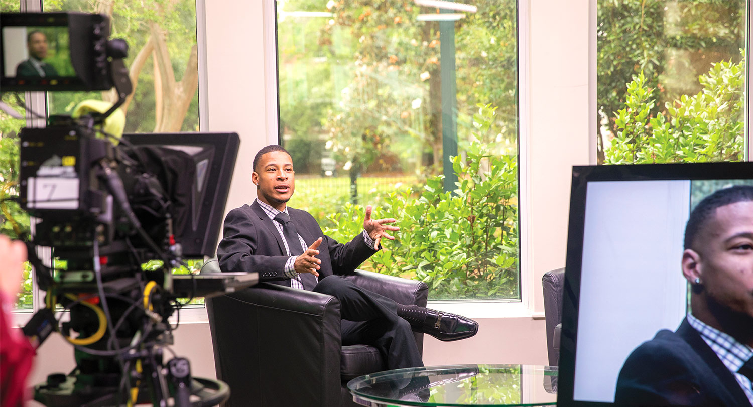 Leland Williams sitting in an armchair gesturing with his hands with a tv camera pointed at him recording an interview.