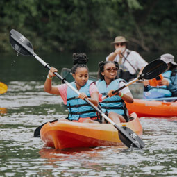 Two students wearing life jackets paddle an orange kayak on a calm river while other kayakers follow behind.