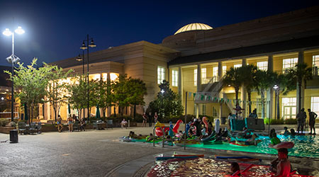 Evening view of the outdoor pool with floats and chairs around it with the fitness center in the background.