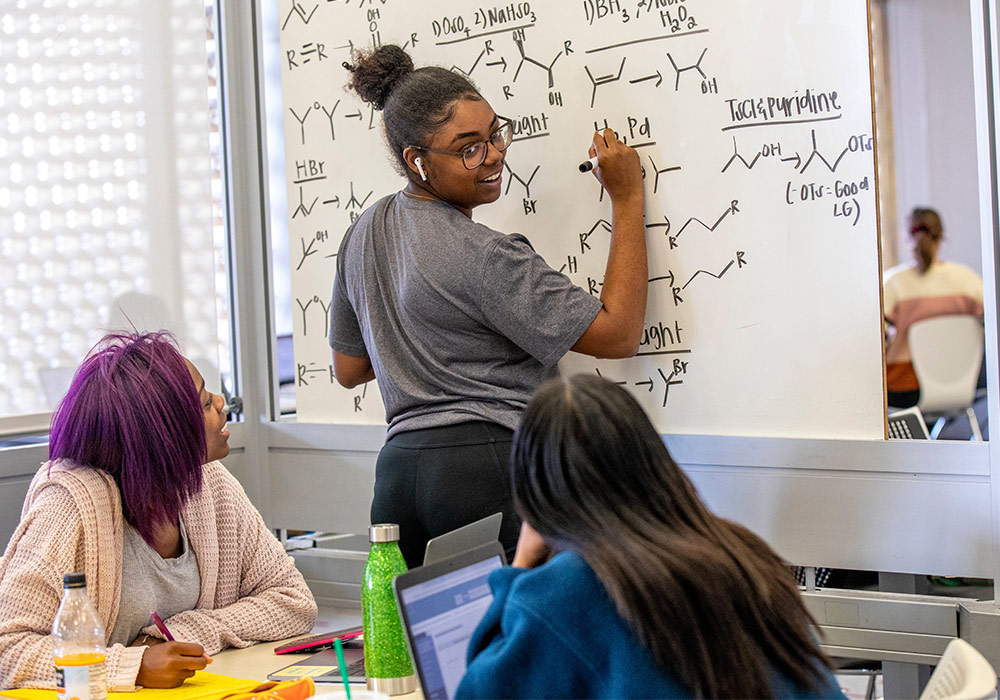 Student writing organic chemistry formulas on a whiteboard turned around looking at two other students sitting at a table.