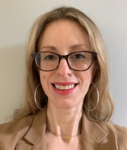 Abbe Schriber has blonde hair and wears gold earrings and a khaki colored blouse. She smiles at the camera in a close-up.