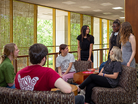 Students gather in the library for a casual discussion. Some are standing and some are sitting.