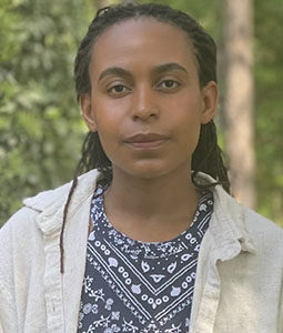 Portrait of a person with dark braided hair, wearing a blue patterned shirt and light outer layer, standing in a wooded area.