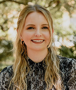 Portrait of a young woman with long wavy blonde hair, wearing a black patterned blouse, smiling outdoors in natural light.