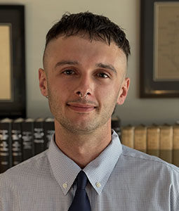 Portrait of a man with short dark hair and a nose ring, wearing a light blue button-up shirt and navy tie, in front of a bookshelf.