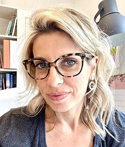 Portrait of a woman with shoulder-length blonde hair wearing tortoiseshell glasses and a grey top, smiling indoors.