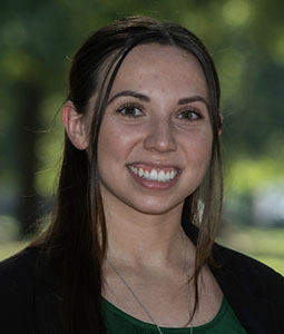 Portrait of a woman with long dark hair, wearing a green top and black blazer, smiling outdoors with greenery in the background.