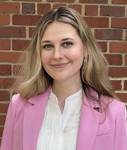 Portrait of a woman with long light brown hair, wearing a pink blazer and white blouse, standing in front of a red brick wall.