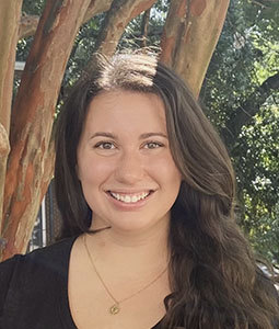 Portrait of a woman with long dark brown hair, wearing a black top and gold necklace, smiling outdoors beside a tree.
