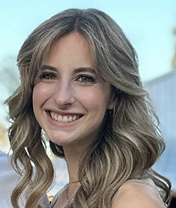 Portrait of a woman with curled blonde hair, wearing a sleeveless top, smiling in front of a blue-toned background.