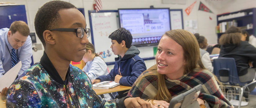 student teacher crouches down to talk with a student in a busy classroom