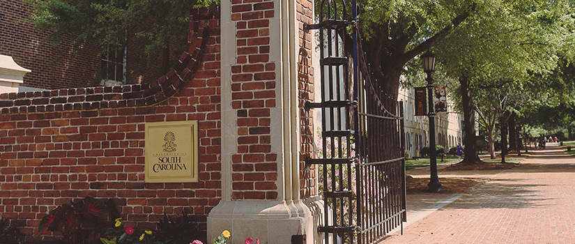 Large brick wall and column with a gold University of South Carolina plaque and the gate open in welcome to the historic Horseshoe area of campus, looking down the brick sidewalk to green space and students walking in the distance..