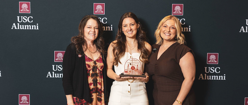 Three women posing with an award in front of a USC banner
