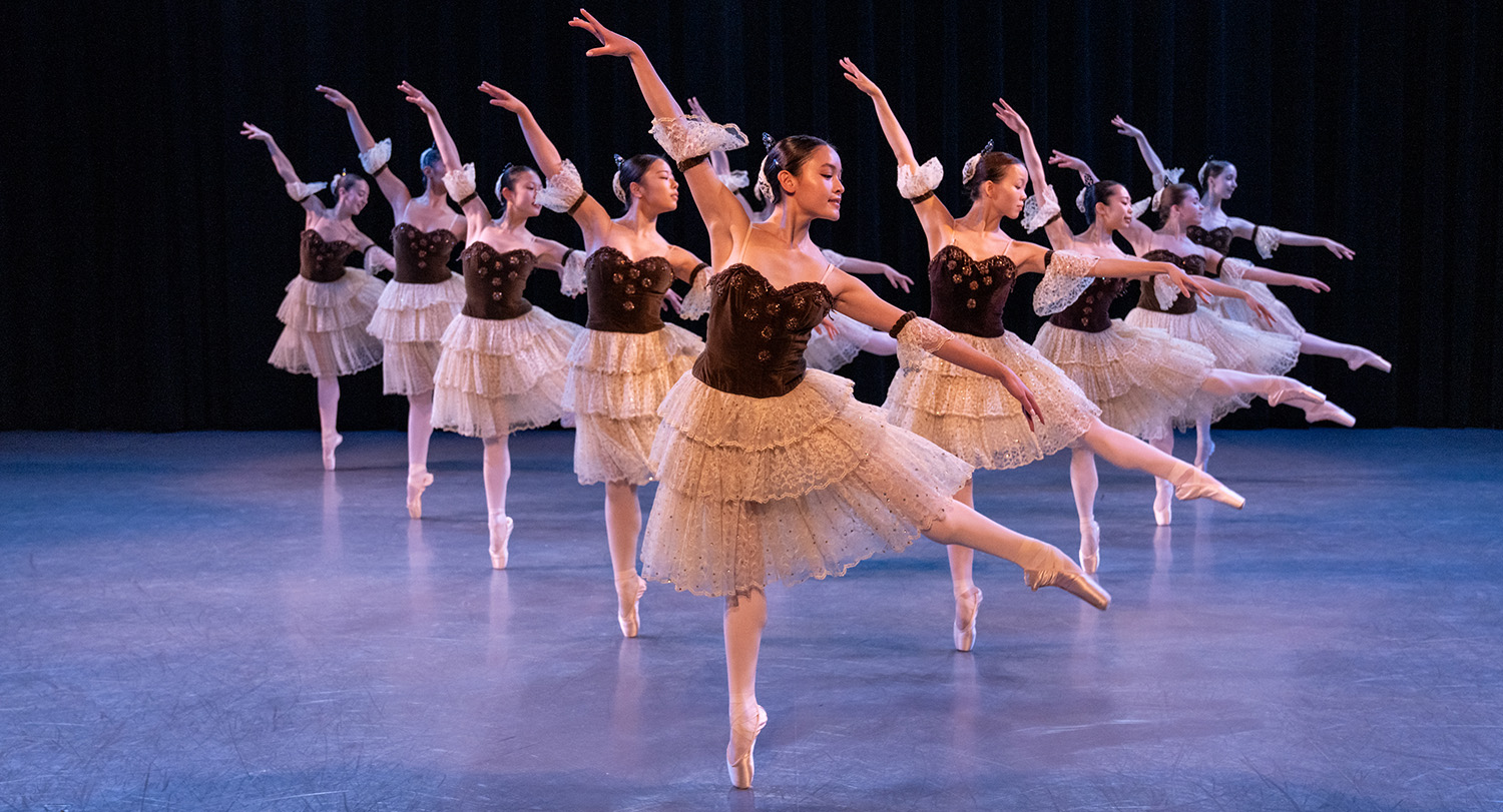 A group of ballet dancers performs on stage in elegant formation, wearing matching brown bodices and layered cream lace tutus. Each dancer is poised on pointe with one leg extended behind and one arm gracefully raised, illuminated against a dark backdrop.