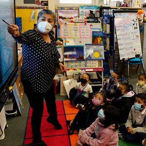 Teacher wearing black and white polka dotted shirt pointing at an electronic board while teaching elementary school students