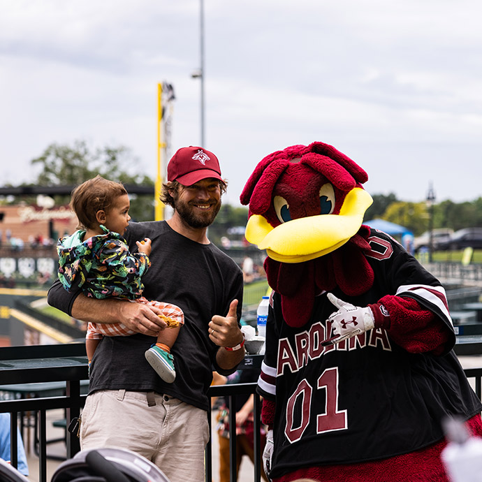 A man holding a small child smiling with Cocky at Segra Park.