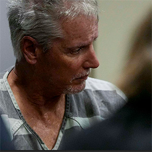 A gray-haired white male in striped prison garb surrounded by people in suits in courtroom setting