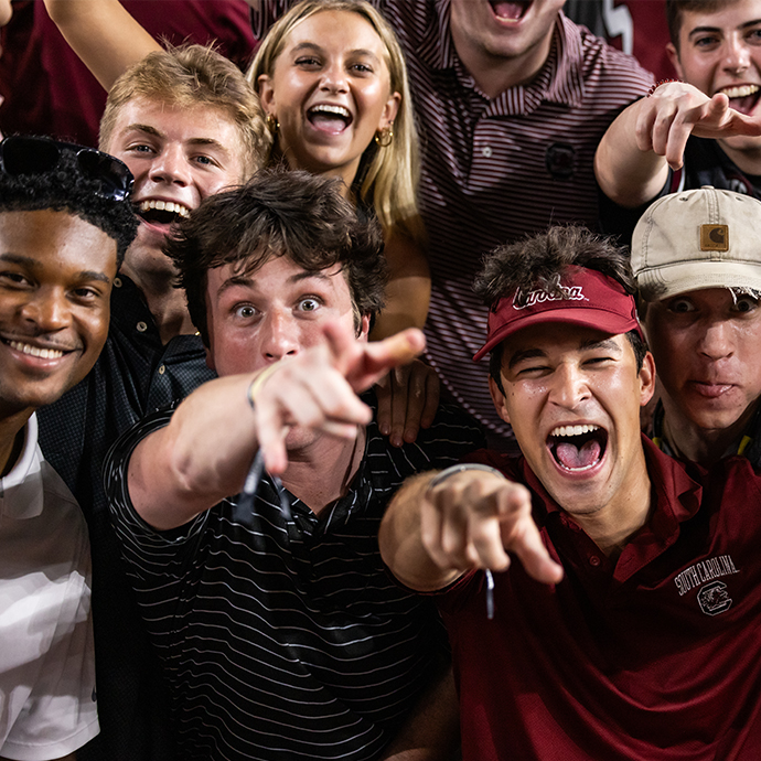 A group of students smiling at a USC football game pointing and smiling.