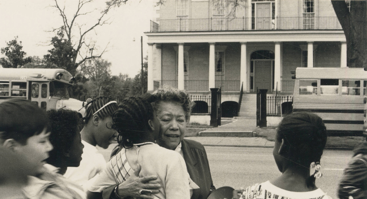 August Baker embrace a child with one arm while greeting others who crowd around her. School buses are visible on the street behind. 