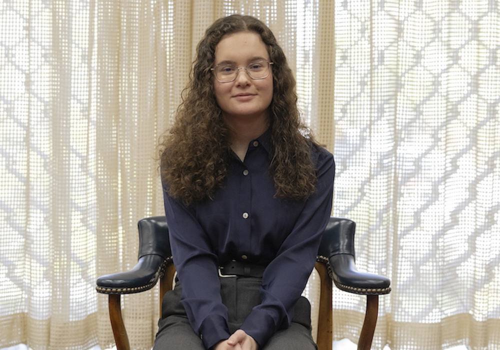 reagan stanton in blue shirt and gray pants sitting in a chair in front of a window